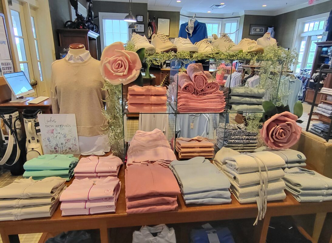 Retail display table in a golf shop featuring neatly folded pastel apparel in pink, green, and neutral tones, accented with decorative flowers, greenery, and a “Ladies new arrivals” sign