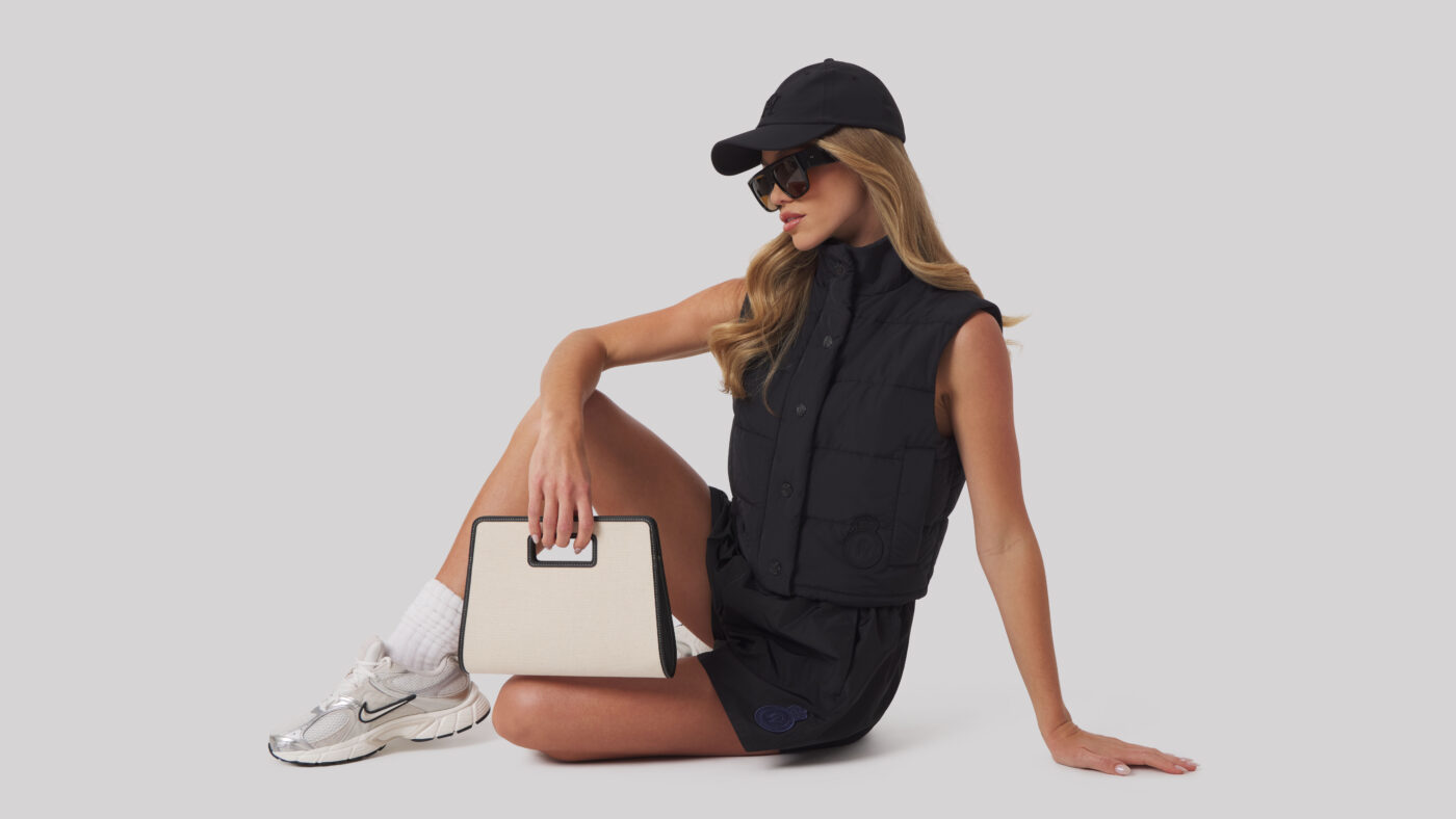 Female model seated against a neutral background wearing a black sleeveless vest, black shorts, and a black cap, holding a structured cream handbag.
