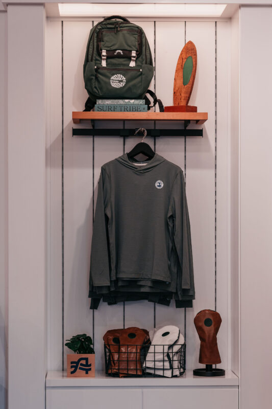 Retail display featuring a Pebble Beach hooded pullover surrounded by surf-inspired accessories, including a backpack, books, wooden décor, and golf headcovers.