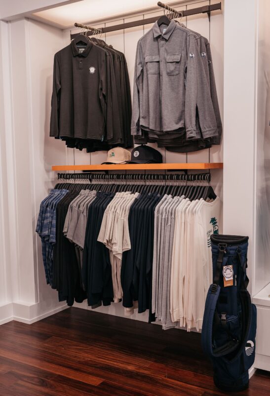 Golf shop apparel display featuring neatly merchandised men’s polos, pullovers, hats, and outerwear in neutral tones, with a golf bag positioned beside the fixture.