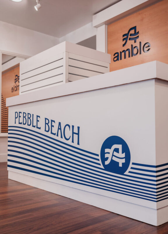 Custom retail checkout counter at Pebble Beach featuring Amble branding with blue wave graphics and wood-paneled logo signage in the background.