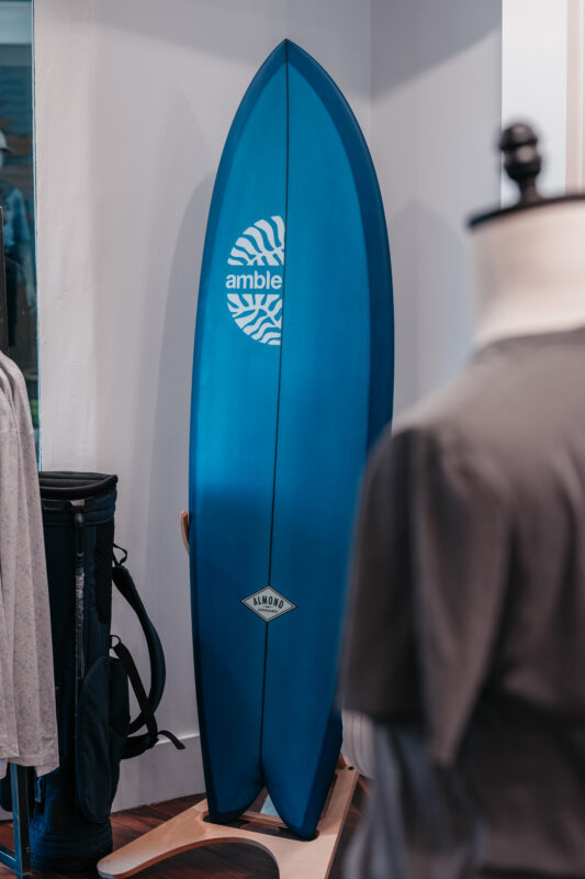 Blue Amble-branded surfboard displayed inside a retail pop-up shop beside golf apparel and accessories.