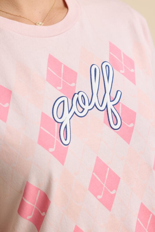 Close-up detail of pale pink golf shirt featuring pink argyle golf club pattern and embroidered blue “golf” script.
