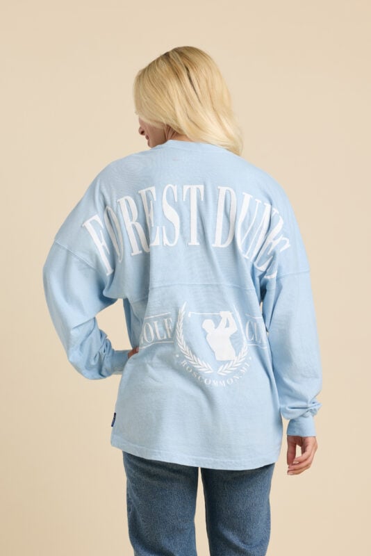 Woman wearing light blue oversized Spirit Jersey-style long sleeve with large white “Forest Dunes” lettering across the back and golf club crest graphic near the hem.