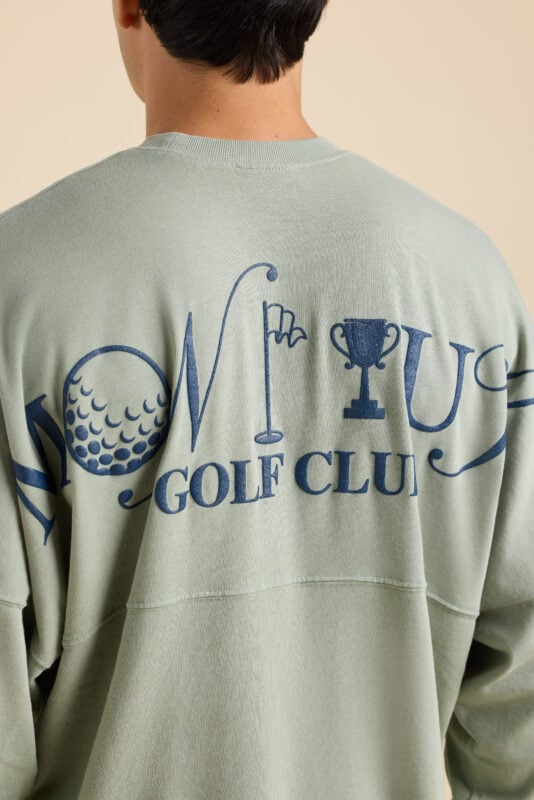 Close-up of sage green sweatshirt back graphic reading “Montauk Golf Club” with golf ball, flagstick, and trophy illustrations.