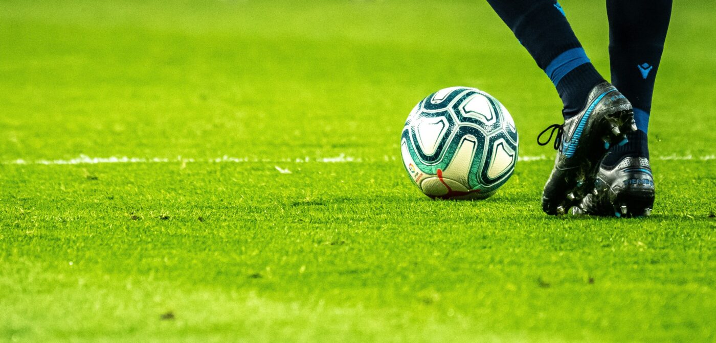 Green soccer field with a man kicking a white and teal soccer ball