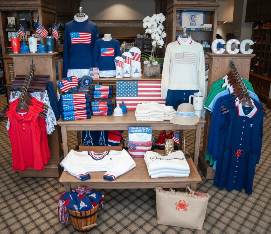 Patriotic golf shop display featuring coordinated red, white, and blue apparel and accessories arranged around an American flag theme. Mannequins showcase navy and white sweaters with flag-inspired graphics, while folded sweaters, polos, dresses, and accessories create layered merchandising throughout the display. A wooden American flag sign serves as the centerpiece, surrounded by miniature U.S. flags, patriotic headcovers, tumblers, and hats. Coordinated apparel racks frame the display with bright red sleeveless tops on one side and blue, green, and white garments on the other. Decorative elements, including illuminated "CCC" letters, white orchids, woven baskets, and a straw tote bag with a red crab motif, add texture and reinforce the club's coastal aesthetic.