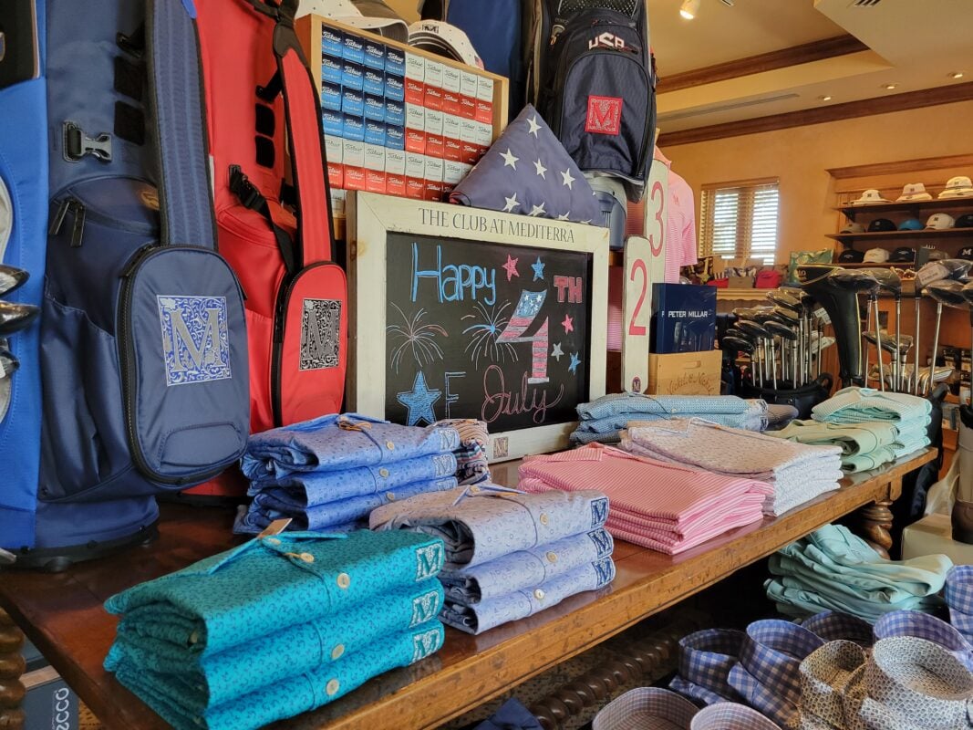 Golf shop display celebrating the Fourth of July with a patriotic mix of merchandise and Americana décor. A rustic wooden table showcases neatly folded polos in shades of blue, pink, white, and mint, arranged around a chalkboard sign reading "Happy 4th of July" with hand-drawn fireworks and stars. Decorative elements include a folded American flag, oversized red numerals, and a wall of red, white, and blue golf ball boxes arranged in a flag-inspired pattern. Monogrammed golf bags in red and navy frame the display, while additional apparel, headwear, and golf equipment fill the surrounding shop. The layered presentation combines patriotic accents with coordinated summer apparel to create an inviting seasonal retail display.