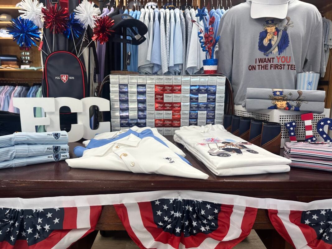 Patriotic golf shop table display featuring red, white, and blue merchandise arranged for America's 250th anniversary. The wooden display table is accented with American flag bunting and showcases folded polos, graphic T-shirts, striped accessories, and leather goods. A gray T-shirt with the phrase "I Want You on the First Tee" and an Uncle Sam illustration serves as the focal point, paired with a coordinating white cap. Decorative elements include red, white, and blue pom-poms, sparkling centerpieces, oversized white "FCC" letters, a wall of color-coordinated golf ball boxes, and freestanding "USA" letters. A navy golf bag embroidered with the club logo and rows of light blue apparel hanging in the background complete the themed presentation.