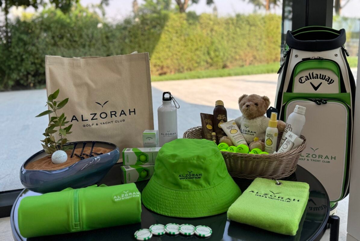 Golf shop wellness display featuring sun protection and sustainable accessories in a coordinated green color palette. Branded items from Al Zorah Golf & Yacht Club include a golf bag, tote bag, bucket hat, towel, reusable water bottles, and golf balls arranged alongside sunscreen products and sunglasses. A woven basket displays skincare essentials and accessories, while natural elements and reusable merchandise reinforce the club's wellness and sustainability focus. Large windows overlooking greenery create a bright, outdoor-inspired setting.