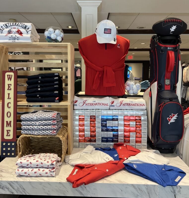 Golf shop display featuring a red, white, and blue merchandise collection celebrating American heritage. A red quarter-zip pullover with a coordinating white cap embroidered with an American flag serves as the centerpiece, flanked by folded polos and patterned apparel in patriotic colors. A navy, red, and white golf bag stands alongside a wall of red, white, and blue golf ball boxes arranged in a flag-inspired pattern. Wooden crate fixtures, woven baskets, and glass bowls filled with golf balls add dimension and texture to the display. Branded apparel from The International Golf Club, including tops with "The International 1901" logo, is showcased alongside a decorative "Welcome" sign with star accents, creating a cohesive and elevated retail presentation.