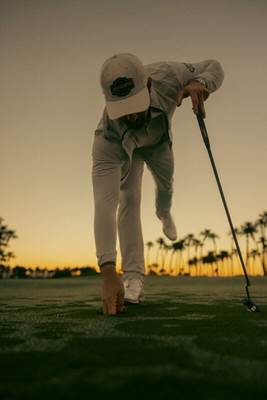 Low-angle photograph of a golfer placing a ball marker on a putting green at dusk. The golfer wears all-white apparel and an Ashworth-branded cap, with palm trees and a sunset sky creating a dramatic backdrop.