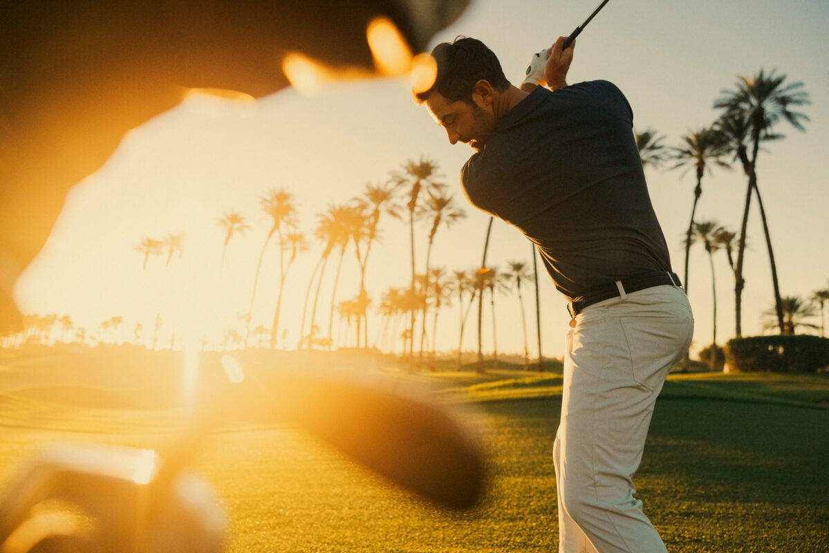 Low-angle photograph of a golfer placing a ball marker on a putting green at dusk. The golfer wears all-white apparel and an Ashworth-branded cap, with palm trees and a sunset sky creating a dramatic backdrop.