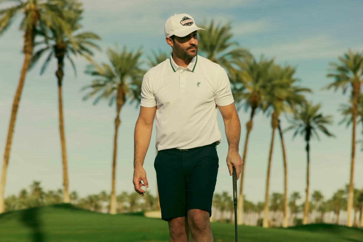 Golfer wearing an Ashworth polo, shorts, and branded cap walks across a palm tree-lined golf course carrying a golf ball and putter. The image highlights Ashworth’s classic golf style with a clean, understated look set against a warm desert course backdrop.