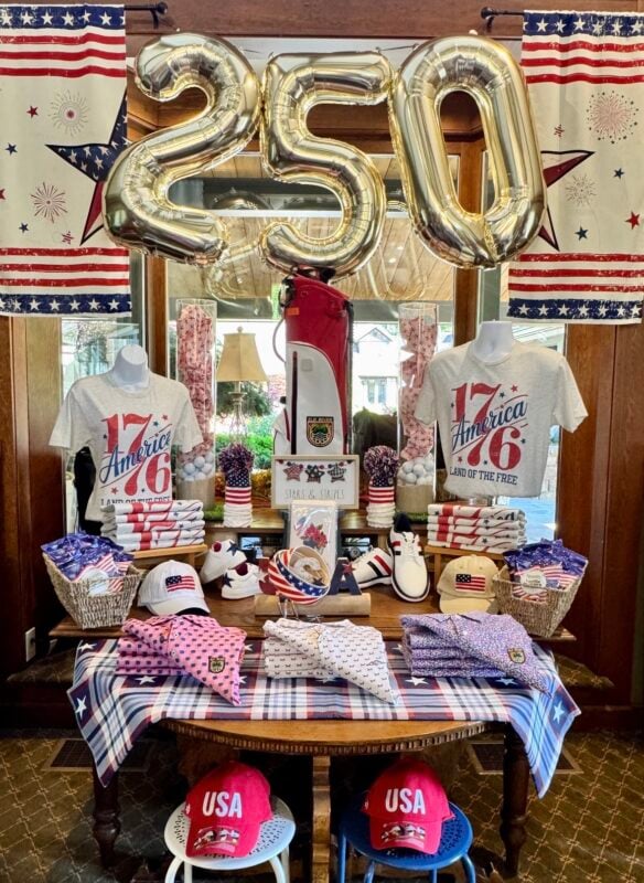 Patriotic golf shop display celebrating America's 250th anniversary, featuring large gold "250" balloons suspended above a red, white, and blue merchandise arrangement. Two mannequins display "1776 America" graphic T-shirts alongside folded apparel, patriotic golf shoes, flag-themed hats, and accessories. A red-and-white golf bag stands at the center, surrounded by decorative golf balls, striped containers, and Americana accents. American flag banners frame the display, while a plaid stars-and-stripes tablecloth and coordinated merchandise create a festive, layered presentation.