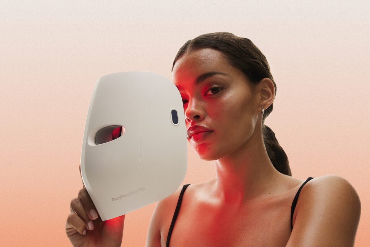 Person holding a Therabody Theraface Mask Glo LED light therapy device beside their face, illuminated with a red light treatment. The image features a soft peach background and highlights the mask's sleek design and skincare-focused wellness technology.
