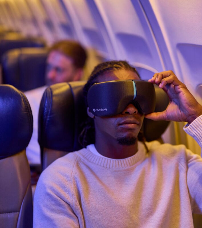 Person relaxing in an airplane seat while using a Therabody smart eye mask during a flight. The traveler wears a light-colored sweater and adjusts the eye mask, which is designed to promote relaxation and recovery while traveling. Soft cabin lighting and blurred passengers in the background create a calm, restful atmosphere.