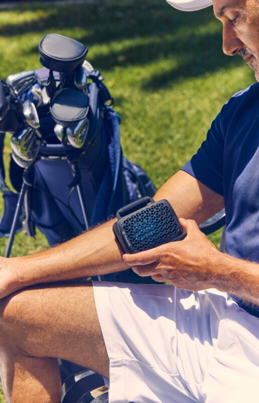 Golfer using a Therabody recovery device while seated beside a golf bag on the course. The portable wellness tool is applied to the forearm, highlighting recovery, circulation, and muscle relief before or after play. The outdoor setting and golf equipment emphasize the growing role of recovery technology in the golf lifestyle.