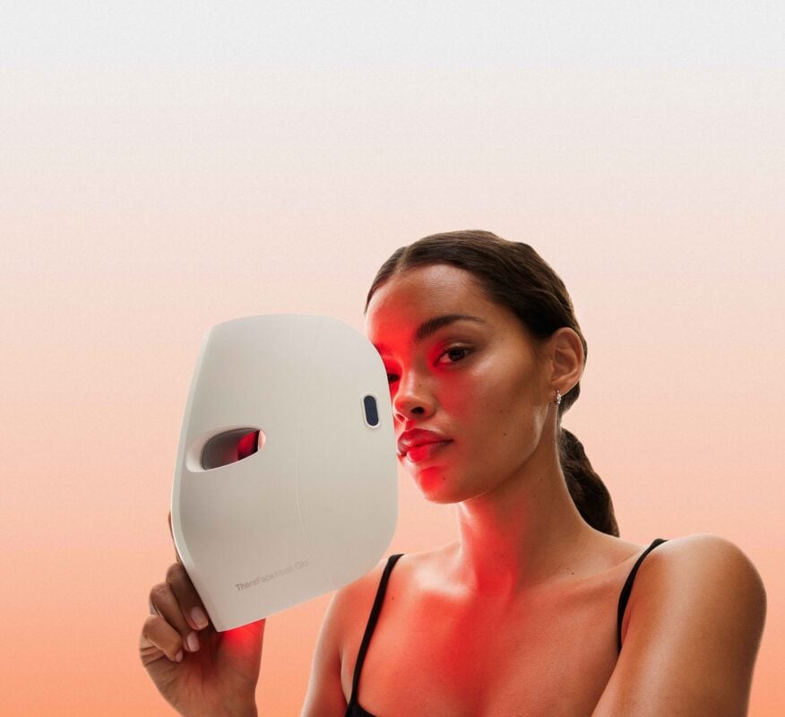 Person holding a Therabody Theraface Mask Glo LED light therapy device beside their face, illuminated with a red light treatment. The image features a soft peach background and highlights the mask's sleek design and skincare-focused wellness technology.
