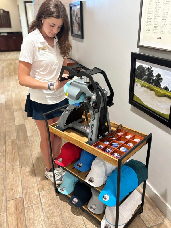 Golf shop staff member operating a hat press machine to customize caps in a retail setting. Standing beside a rolling display cart, the employee wears a white polo, navy skirt, and athletic shoes while applying a design to a light blue cap. The cart features neatly organized hats in a variety of colors, along with a tray of interchangeable patches and emblems for personalization. Framed golf artwork lines the hallway wall, highlighting an interactive customization experience within the club's golf shop.