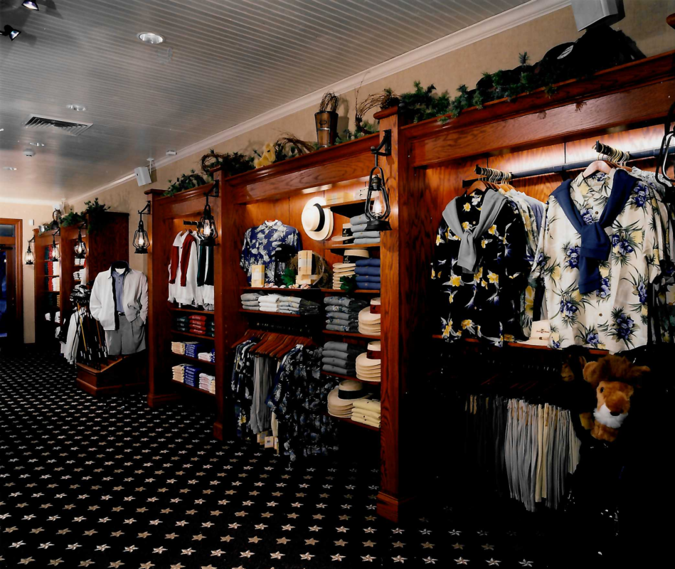 Golf shop apparel displays built into rich wood wall fixtures, featuring coordinated men's merchandise including floral prints, layered knitwear, polos, hats, and accessories. Warm accent lighting, decorative lanterns, and themed visual merchandising create a refined, country club-inspired retail environment with strong color coordination and vertical presentation.