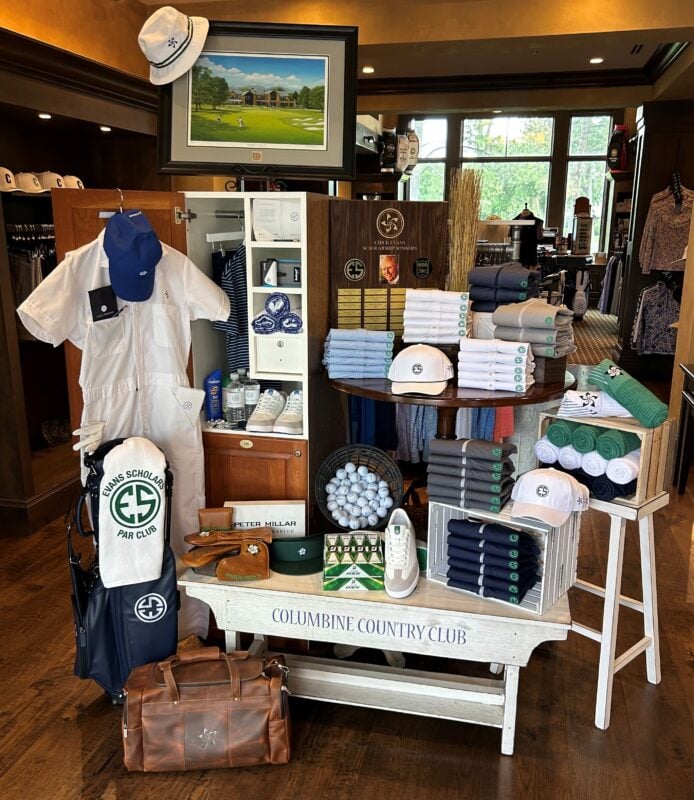 Golf shop display celebrating caddie culture and club traditions, featuring coordinated apparel, accessories, and gifts in the club's signature colors. A mannequin dressed in a caddie-style coverall stands beside a branded golf bag, while folded shirts, hats, towels, and golf balls are arranged on tables and wooden crates. A scholarship recognition display and framed course artwork reinforce the club's heritage, creating a merchandise presentation that highlights both community and tradition.