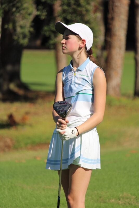 Young golfer wearing a white cap and sleeveless Calleigh’s Way dress stands on the course holding a driver while watching play ahead.