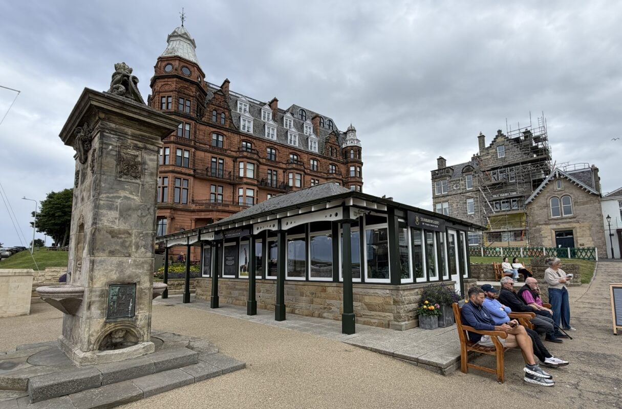 Street-level photo in St Andrews showing the historic Royal and Ancient Golf Club area, with ornate stone and brick architecture, a golf shop, and visitors seated outside beneath an overcast sky.