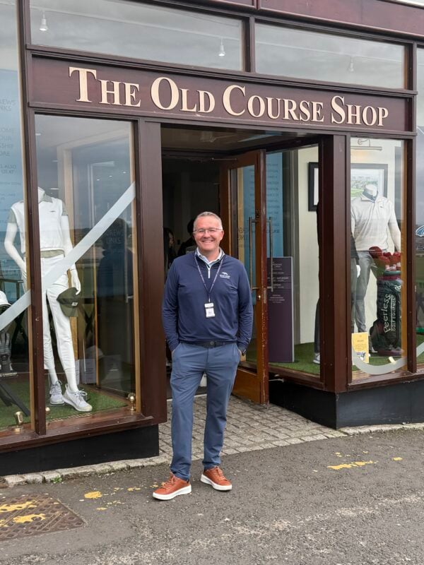 Photo of a staff member standing outside The Old Course Shop at St Andrews Links, with windows displaying official golf apparel and merchandise.