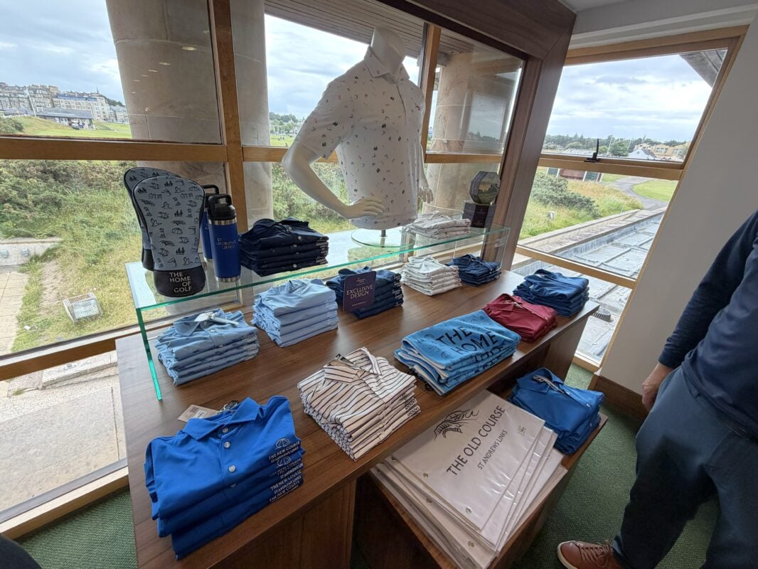 Photo of an Old Course Shop apparel display featuring folded St Andrews Links polos and shirts, a mannequin, and branded golf accessories beside large windows overlooking the course.