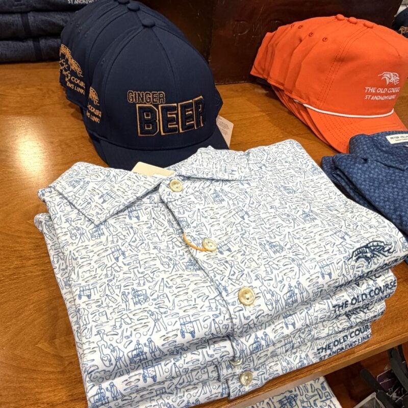 Photo of folded Old Course St Andrews golf shop merchandise, featuring patterned polos alongside navy “Ginger Beer” and orange branded caps.