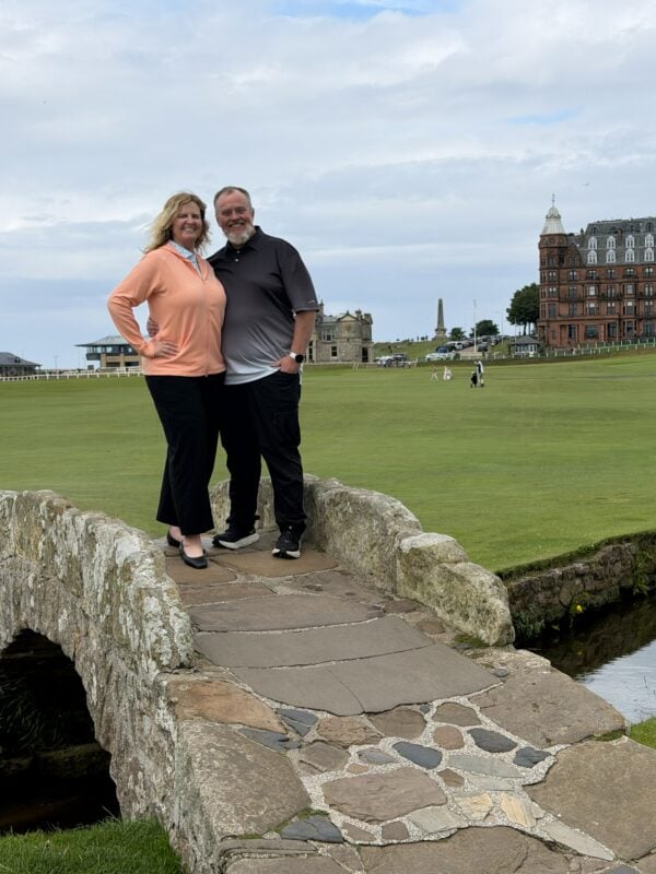 Photo of a Jennifer Morton and Ken Morton Jr. standing on the historic Swilcan Bridge at the Old Course at St Andrews, with the fairway and iconic clubhouse architecture in the background.