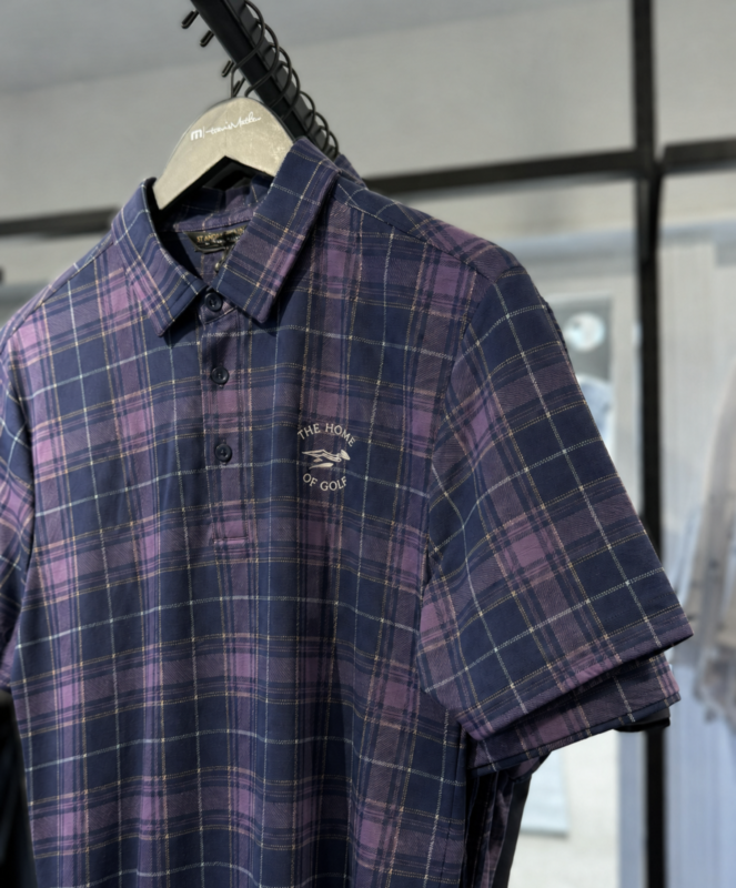 Close-up photo of a purple and navy tartan golf polo hanging in a pro shop, featuring an embroidered “The Home of Golf” logo on the chest.