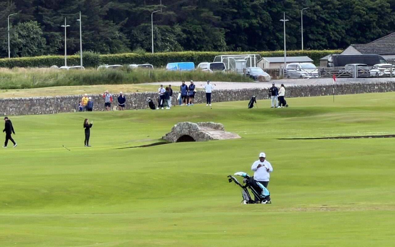 Wide view of golfers and visitors on the Old Course at St Andrews, with the historic Swilcan Bridge centered on the fairway.