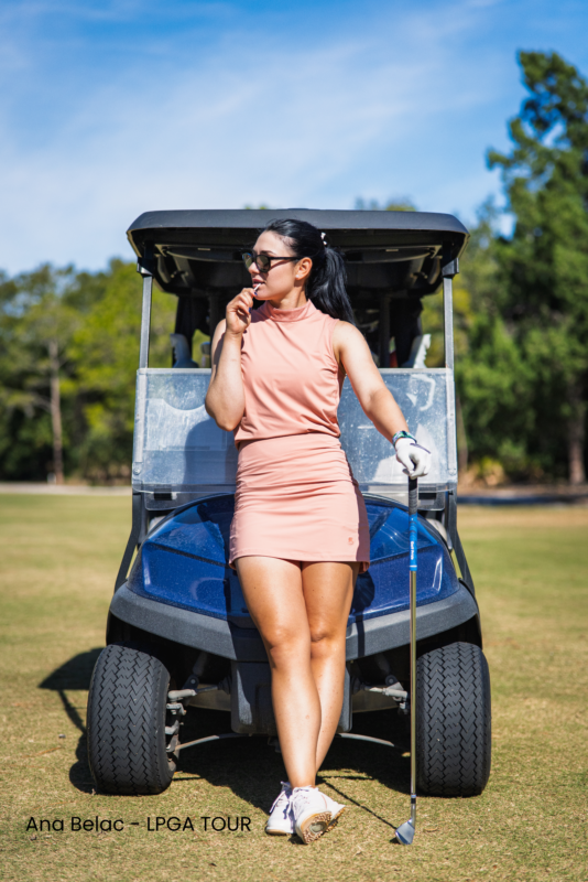 Lifestyle image of LPGA Tour professional Ana Belac wearing women's golf apparel on a golf course. Ana leans casually against the front of a blue golf cart while wearing a blush pink sleeveless mock-neck golf top with a coordinating golf skirt, white golf glove, white golf shoes, and dark sunglasses. She rests one hand on a golf club and looks off into the distance with a relaxed expression. The image is set on a sunny golf course with green fairways, trees, and a blue sky in the background, showcasing stylish, performance-ready women's golf apparel in an authentic on-course setting.