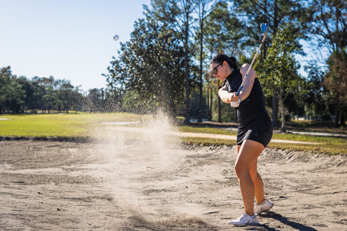 Action photograph of LPGA Tour professional Ana Belac wearing women's golf apparel while playing from a sand bunker on a golf course. Ana is captured mid-swing as she strikes a golf ball from the bunker, sending a spray of sand into the air. She wears a black sleeveless mock-neck golf top, coordinating black golf skort, white golf glove, white golf shoes, and dark sunglasses. Trees, fairway, and a clear blue sky form the background, while the dynamic motion highlights the performance, flexibility, and athletic design of the women's golf apparel in authentic on-course play.
