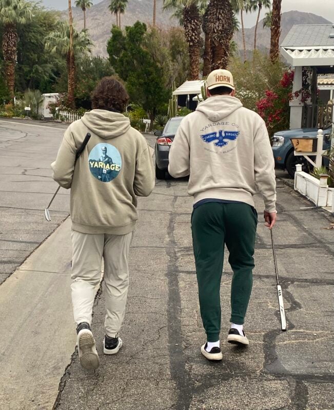 Two people walk down a palm-lined street carrying golf clubs and wearing Yardage Company hoodies with large branded graphics on the back. Mountains rise in the background.