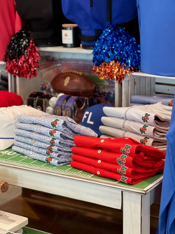 Golf shop display featuring neatly folded red, white, and patterned Florida Gators polos, with football-themed décor, pom-poms, and a football in the background.