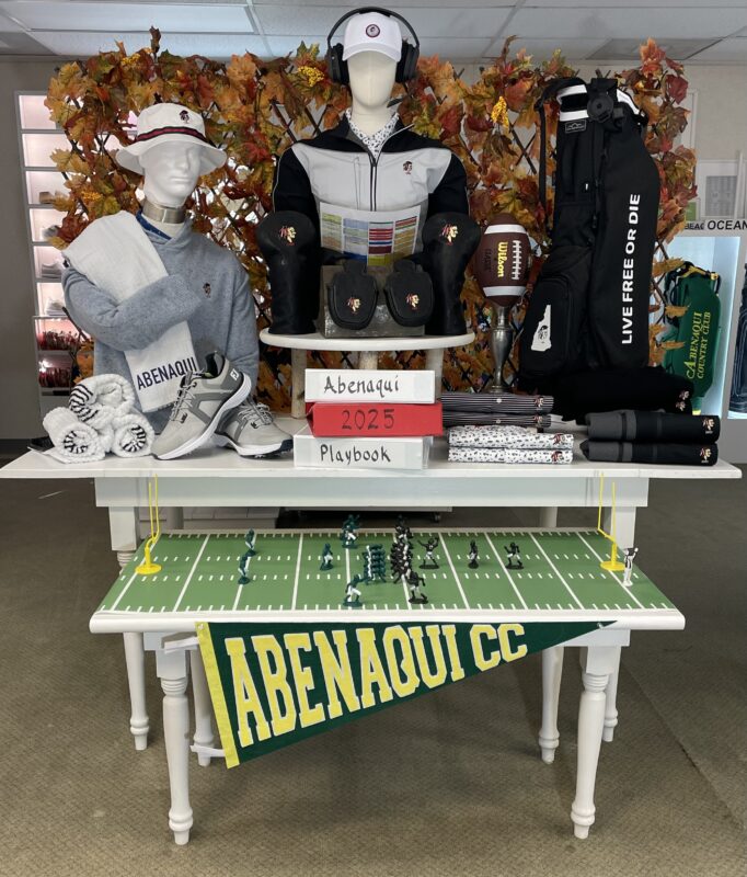 Football-themed golf shop display at Abenaqui Country Club featuring mannequins in club apparel, golf accessories, a football, fall foliage, and a miniature football field with players and goalposts beneath an “Abenaqui CC” pennant.