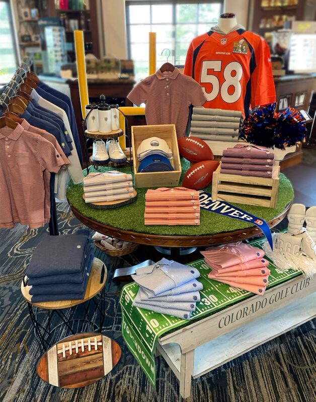 Football-themed display at Colorado Golf Club featuring folded club apparel, footballs, hats, golf shoes, Denver Broncos décor, orange-and-blue pom-poms, and a mannequin wearing a Broncos No. 58 jersey.