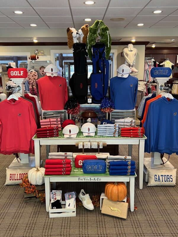 Football-themed golf shop display featuring red and blue collegiate apparel, hats, golf bags, pom-poms, football décor, and Bulldogs and Gators merchandise arranged around a central table.