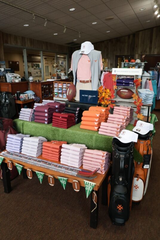 College football-themed golf shop display featuring neatly folded Texas, Texas A&M, Texas Tech, and TCU apparel, footballs, golf bags, fall leaves, and yard-line decorations.
