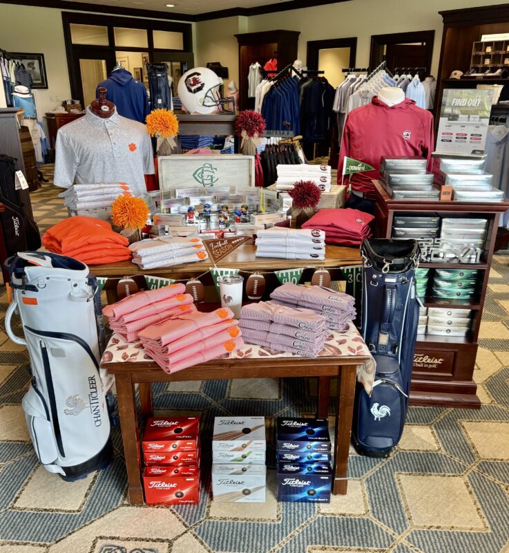 College football-themed golf shop display featuring Clemson and South Carolina apparel, football helmets and décor, golf bags, pom-poms, and stacks of Titleist golf balls.