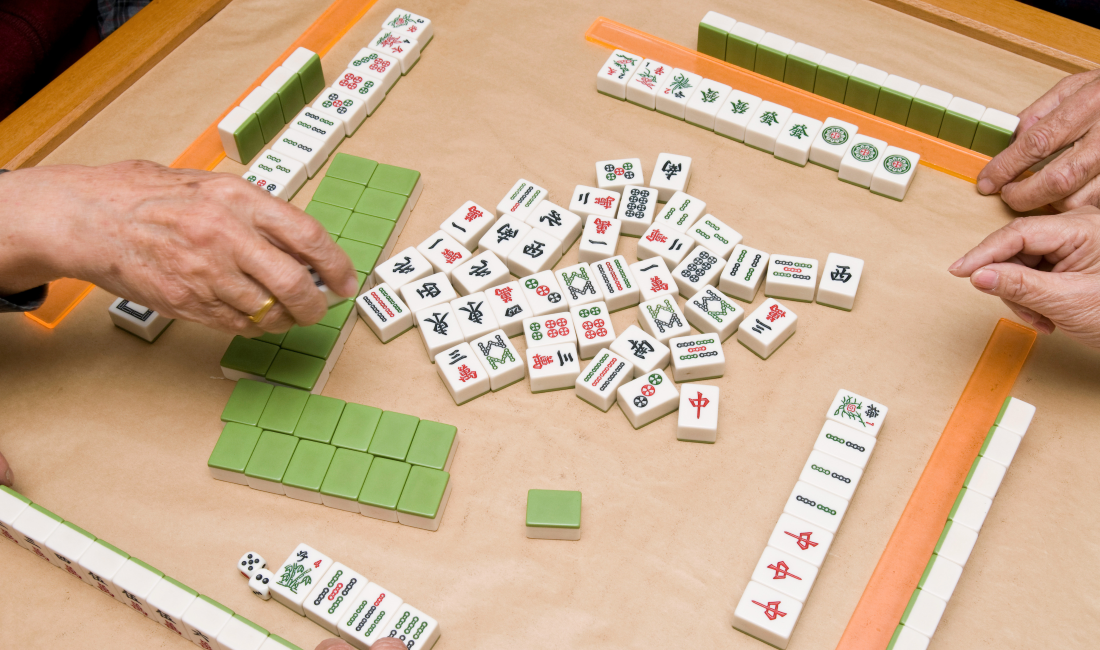 Photo of people playing mahjong, with hands reaching toward green-and-white tiles arranged on racks and scattered across the game table.