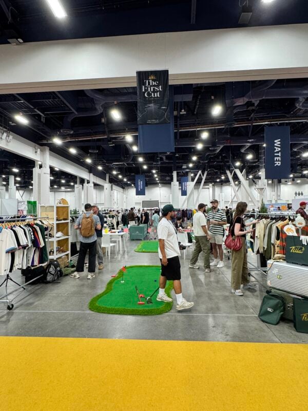 Photo of a busy golf retail trade show floor with attendees browsing apparel displays and putting greens set up throughout the exhibit space.