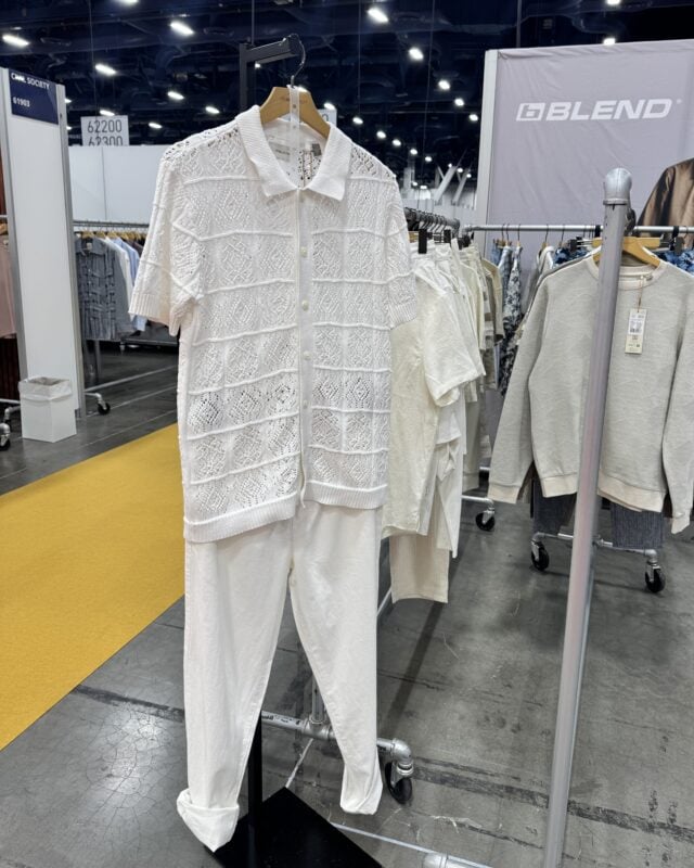 Photo of an all-white outfit featuring a textured knit button-up shirt and pants displayed at a retail trade show.