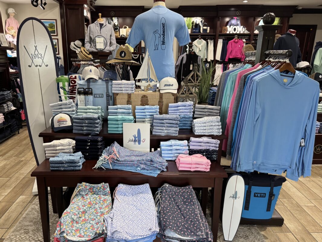 Photograph of a golf shop merchandise display at Woodmont Country Club in Rockville, Maryland, featuring neatly folded johnnie-O apparel, patterned shorts, hats, YETI coolers, and nautical-themed accessories. A mannequin wearing a blue johnnie-O shirt stands above the display, with additional apparel hanging on surrounding racks.