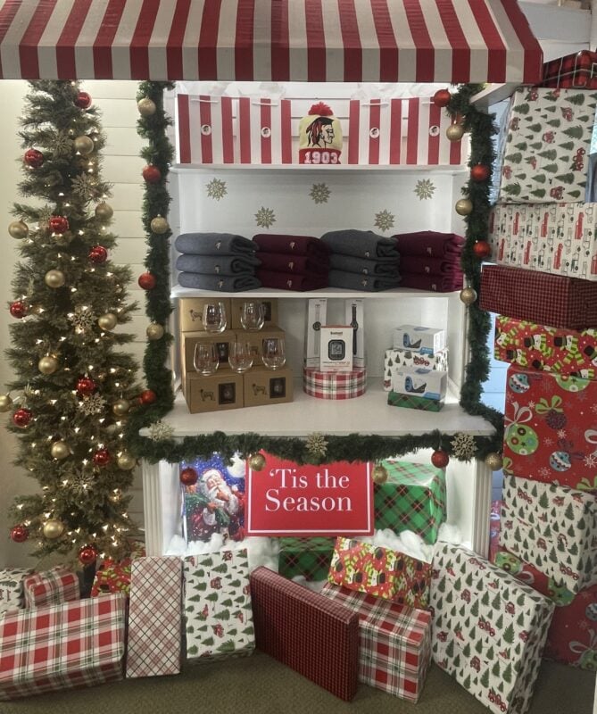 Christmas golf shop display at Abenaqui Country Club featuring a red-and-white striped awning, decorated Christmas tree, garland, folded apparel, gift items, and wrapped presents surrounding a “‘Tis the Season” sign.