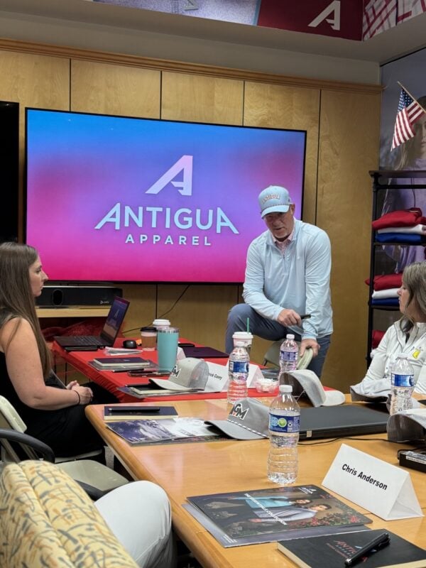 Photo from an AGM Regional Event at Antigua Apparel, showing a presenter in Antigua-branded apparel speaking with attendees seated around a conference table. A large screen displays the Antigua Apparel logo, with AGM hats and event materials arranged on the tables.