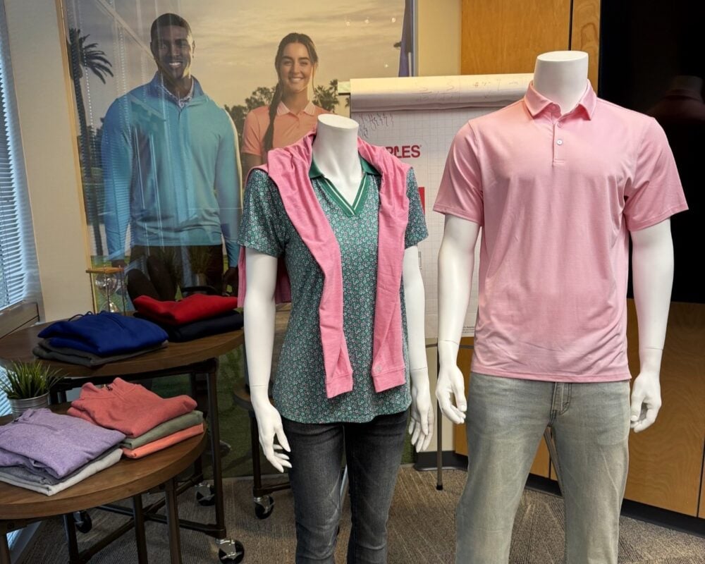 Photo from an AGM Regional Event at Antigua Apparel, showing two mannequins dressed in men's and women's golf apparel. One wears a green patterned polo with a pink sweater draped over the shoulders, while the other wears a pink polo with gray pants. Folded apparel and an Antigua promotional poster are displayed in the background.
